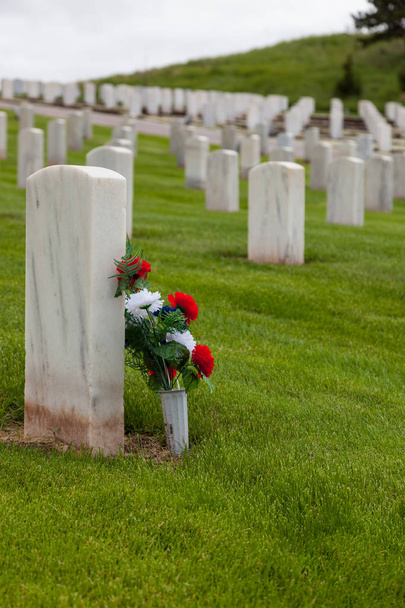 Um grupo de flores vermelhas brancas e azuis encostadas a uma lápide de mármore em um cemitério militar com grama vibrante de primavera em uma colina
. - Foto, Imagem