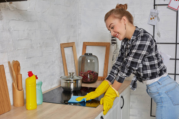Blonde woman in protective gloves with rag cleaning electric stove at home kitchen. Girl washing black shiny surface of kitchen top - Photo, Image