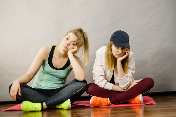 Two attractive women wearing sports clothes sitting on exercise mat being bored or tired after hard workout. - 写真・画像