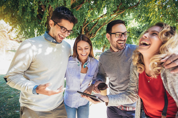 Cuatro felices jóvenes amigos sonrientes caminando al aire libre en el parque sosteniendo la tableta digital
 - Foto, Imagen