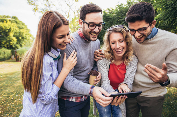 Cuatro felices jóvenes amigos sonrientes caminando al aire libre en el parque sosteniendo la tableta digital
 - Foto, Imagen