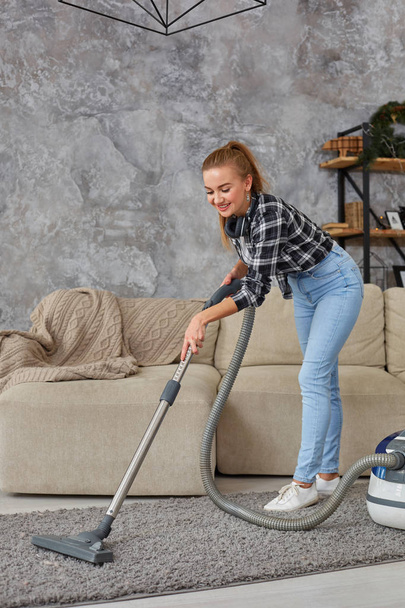 Joven mujer sonriente aspiradora limpiando la alfombra en la sala de estar, interior escandinavo moderno. Hogar, concepto de limpieza
 - Foto, imagen
