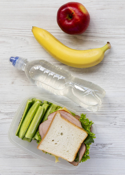 Γεννούν υγιή σχολικό κιβώτιο μεσημεριανό γεύμα με σάντουιτς, φρούτα και ένα μπουκάλι νερό σε λευκή ξύλινη επιφάνεια, επίπεδη. Από πάνω, κάτοψη, γενικά. - Φωτογραφία, εικόνα