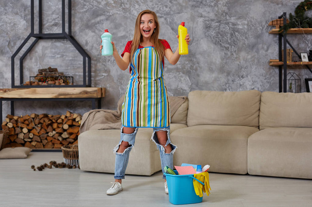 Женщина сравнивает две бутылки стирального порошка в доме уборки со многими предметами домашнего обихода в синем ведре в гостиной. Работа на дому
. - Фото, изображение