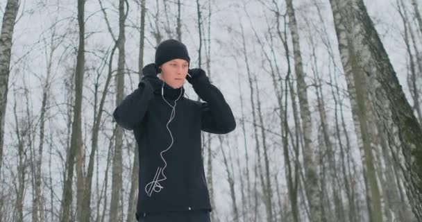 Mladý muž na ráno jog drží sluchátka v jeho rukou a vloží ho do jeho uší před spuštěním v parku. Zimní běh. Školení aplikace a používat moderní pomůcky. Poslouchat hudbu a zároveň - Záběry, video