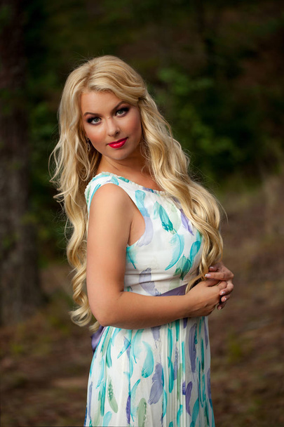 Young beautiful blonde woman in white dress with blue and green flowers posing in forest - Photo, image
