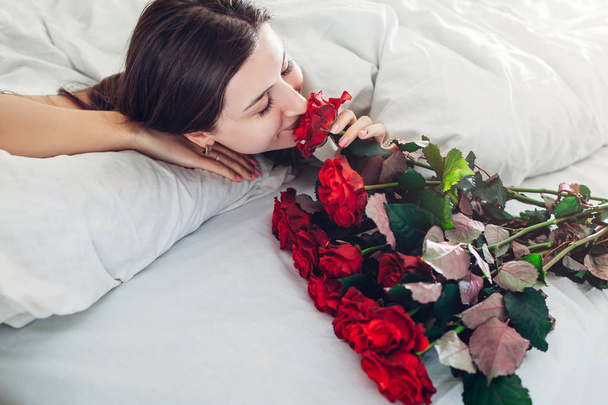 Young woman found bouquet of roses in bed. Happy girl smelling flowers. Women's day surprise - Фото, изображение