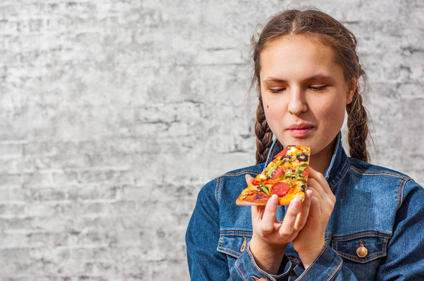 portrait of young teenager brunette girl with long hair eating slice of pizza on gray wall background with copy space. - Fotó, kép