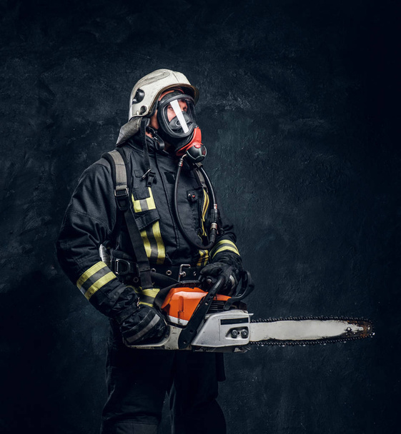 Portrait d'un pompier portant un casque de sécurité et un masque à oxygène tenant une tronçonneuse. Studio photo contre un mur sombre texturé
 - Photo, image