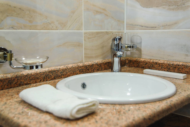 Bathroom. The shell is white. Fold the white towel into a pile. Marble top. The walls are tiled. The interior of the bathroom. Sink mixer. - Фото, изображение