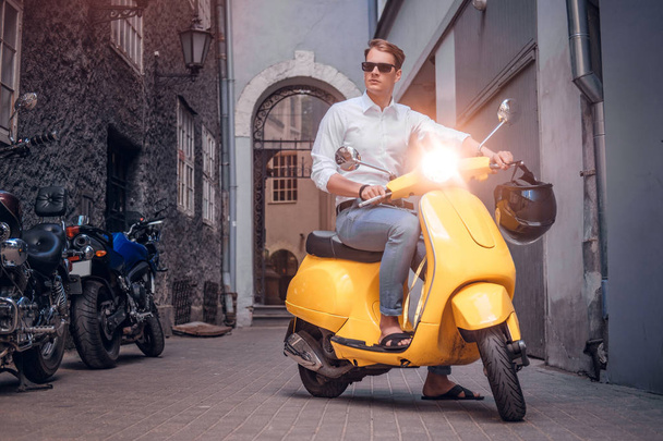 Handsome man wearing sunglasses riding on vintage Italian scooter in the old narrow street of Europe - Foto, afbeelding