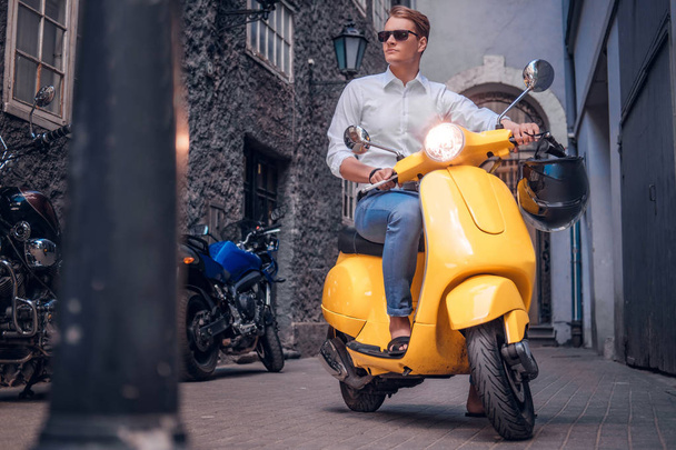 Komea mies, jolla on aurinkolasit ja joka ratsastaa vintage-italialaisella skootterilla Euroopan kapealla kadulla
 - Valokuva, kuva
