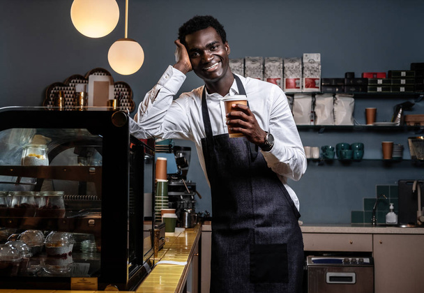 Cansado barista africano sosteniendo una taza con café mientras se apoya en un mostrador en una cafetería y mirando a una cámara con una mirada feliz
 . - Foto, Imagen