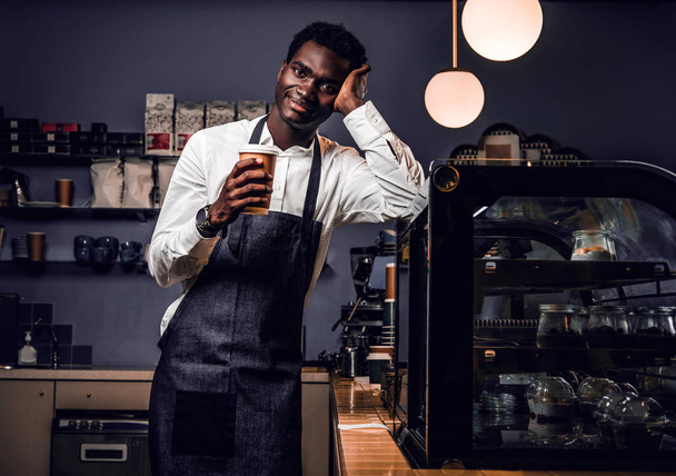 Müder Barista mit einer Tasse Kaffee, während er sich in einem Coffeeshop an einen Tresen lehnt und mit glücklichem Blick in eine Kamera blickt  - Foto, Bild