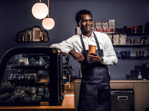 African barista holding a cup with coffee while leaning on a counter in a coffee shop - Zdjęcie, obraz