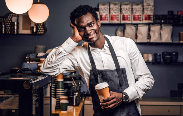 Cansado barista sosteniendo una taza con café mientras se apoya en un mostrador en una cafetería y mirando a una cámara con una mirada feliz
  - Foto, Imagen