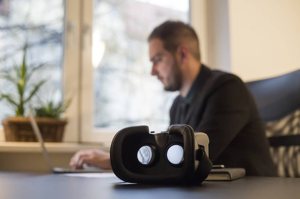 бизнесмен работает в офисе очки виртуальной реальности ждет его у окна черный стол с ноутбуком бумаги телефон VR
 - Фото, изображение