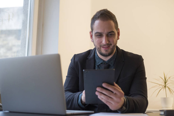 homem de negócios trabalhando no laptop no escritório tomar notas em tablet por janela mesa preta com notebook papéis telefone
 - Foto, Imagem