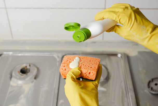 Dvě ruce, použití tekutého mýdla z láhve čističi na oranžová houba. Vytlačení detergent na oranžová houba na pozadí plynový sporák. - Fotografie, Obrázek
