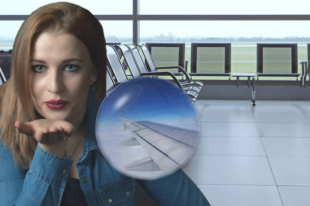 Sensual young woman is sending a kiss with a open palm at the airport terminal. The image of the wing of the flying airplane is in a transparent ball. - Zdjęcie, obraz