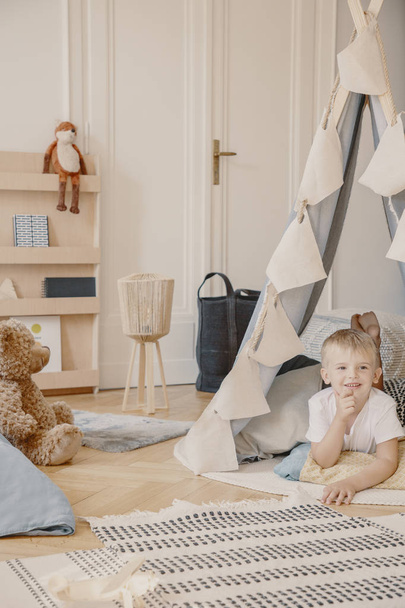 Smiling boy in tent in bright playroom interior with plush toys and patterned rug. Real photo - Foto, Imagen