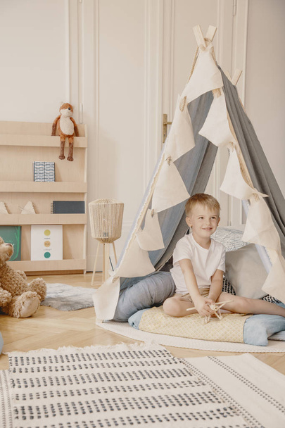 Happy child in tent and rug in white playroom interior with plush toy on wooden shelf. Real photo - Photo, Image