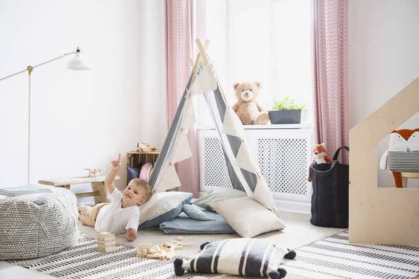 Smiling kid on the floor and plush toy in bedroom interior with pouf and tent next to lamp. Real photo - Foto, Imagen