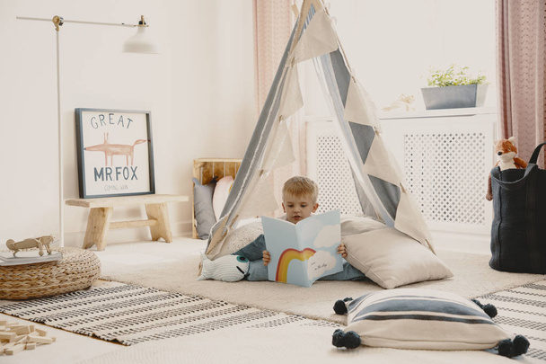 Bambino carino che legge un libro, sdraiato sui cuscini in elegante tenda scandinava in sala giochi
 - Foto, immagini