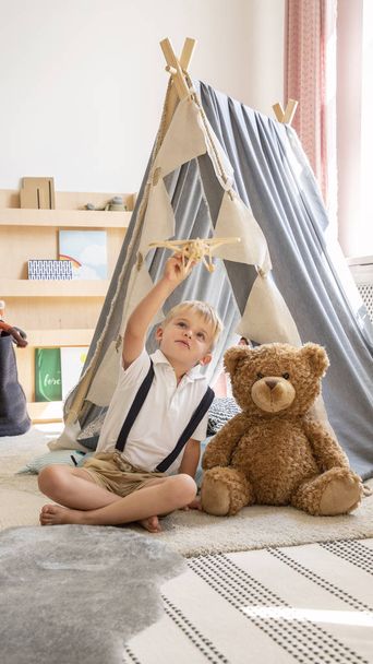 Cute little blonde boy sitting next to teddy bear in stylish scandinavian kid's playroom - Foto, afbeelding