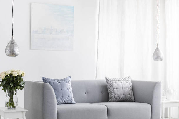 Pillows on grey sofa in white living room interior with lamps, flowers and poster. Real photo - Φωτογραφία, εικόνα