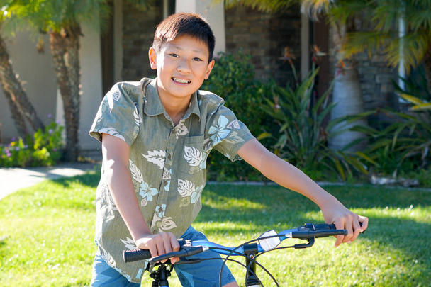 Ο νεαρός Ασίας αγόρι με το ποδήλατό του μπροστά από το σπίτι. Νέοι έφηβος αγόρι στο ποδήλατό του, χαμογελώντας και δείχνοντας του ορθοδοντικά σιδεράκια για τα δόντια του. - Φωτογραφία, εικόνα