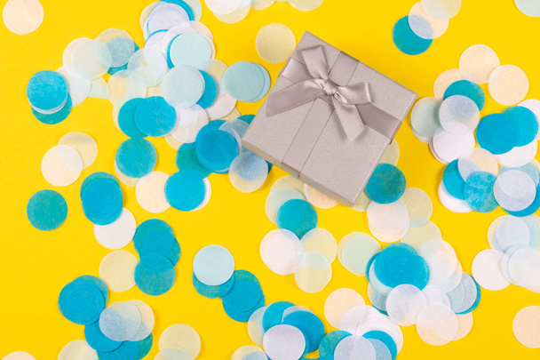 kleine niedliche Geschenkschachtel mit glänzender Satinschleife auf gelbem Hintergrund mit buntem Konfetti. flacher Lagestil. Ferien- und Festkonzept. - Foto, Bild