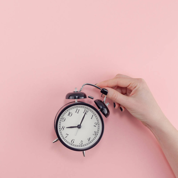Creative flat lay morning time emotions concept top view of woman hand holding black vintage alarm clock pastel millennial pink color paper background with copy space minimal style square template - 写真・画像