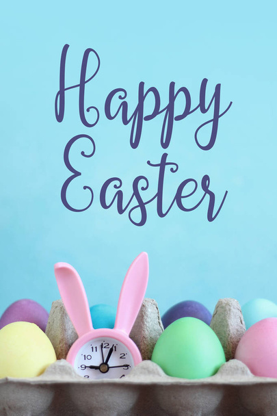 Multicolored Easter eggs and pink bunny alarm clock on the blue background. Happy Easter card - Foto, imagen