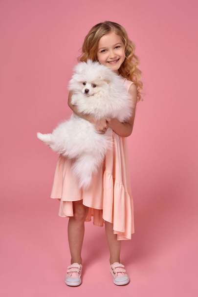 Niña con el pelo rubio rizado, en un vestido rosa juega con su perro
 - Foto, imagen