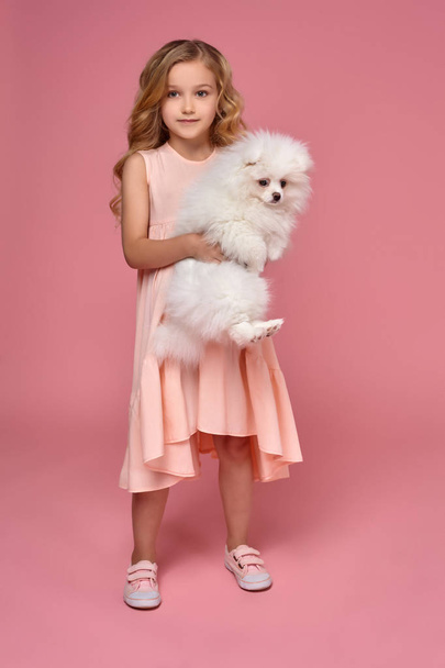 kleines Mädchen mit blonden lockigen Haaren, im rosa Kleid spielt mit ihrem Hund - Foto, Bild