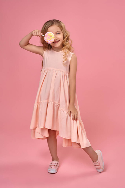 kleines Mädchen mit blonden lockigen Haaren, in einem rosa Kleid posiert mit einem Bonbon - Foto, Bild