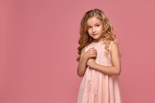 Petite fille aux cheveux bouclés blonds, dans une robe rose pose avec un bonbon
 - Photo, image