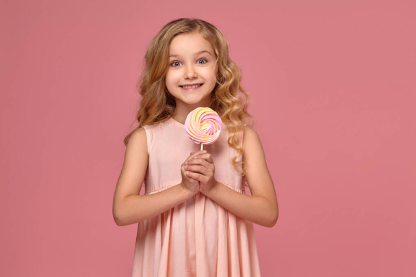 Petite fille aux cheveux bouclés blonds, dans une robe rose pose avec un bonbon
 - Photo, image