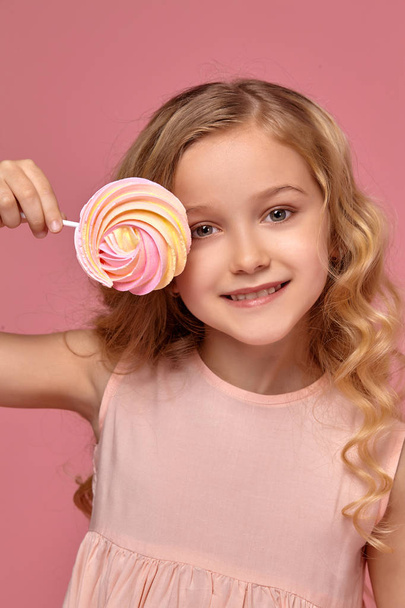 Μικρό κορίτσι με ξανθά σγουρά μαλλιά, σε ένα ροζ φόρεμα ποζάρει με καραμέλα - Φωτογραφία, εικόνα