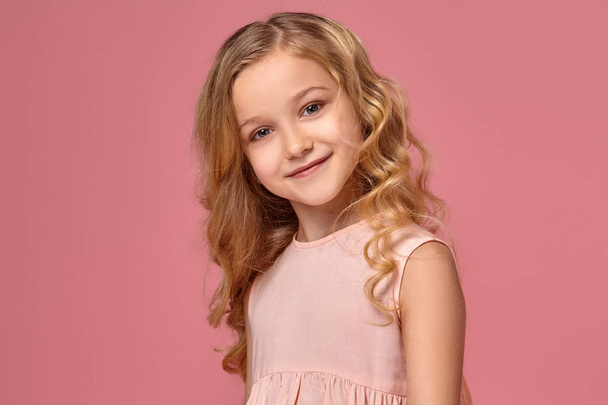 Little girl with a blond curly hair, in a pink dress is posing for the camera - Фото, изображение