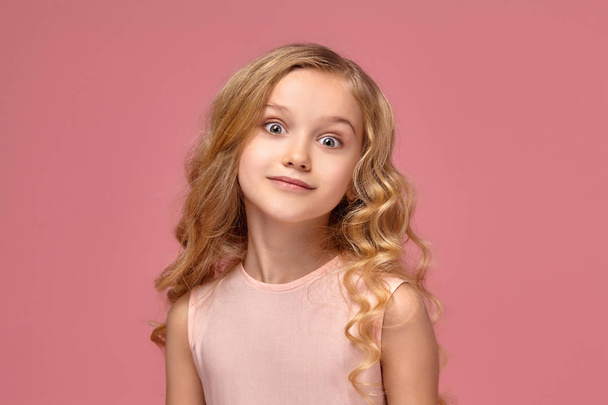 Malá dívka s kudrnatými blond vlasy, v růžových šatech pózuje pro kamery - Fotografie, Obrázek