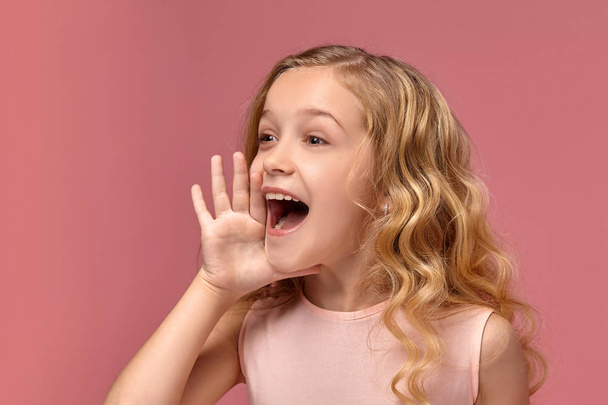 Little girl with a blond curly hair, in a pink dress is posing for the camera - Photo, Image