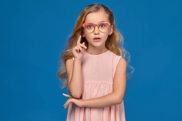 Modna Dziewczynka w różowej sukience i okulary stoi na niebieskim tle. - Zdjęcie, obraz