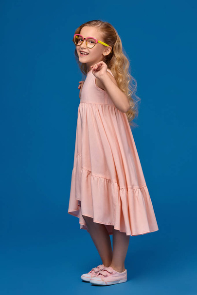 Fashionable little girl in a pink dress and glasses is standing on a blue background. - Foto, Bild