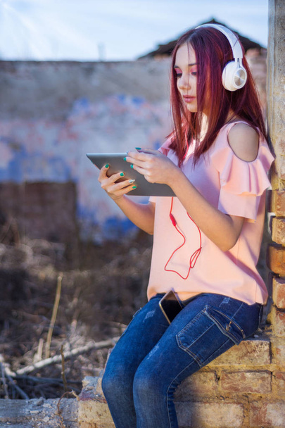 Linda pelirroja dama con tableta digital escuchando música en auriculares en ruinas de la pared de ladrillos rojos de la casa retro en la puesta del sol. Chica encantadora con auriculares blancos disfruta en la vida. Primer plano, enfoque selectivo
 - Foto, imagen