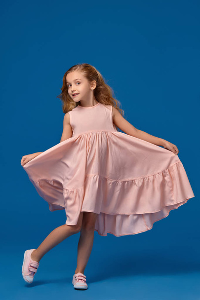 hübsches kleines Mädchen in rosa Kleid, mit lockigem Haar schwenkt ihr Kleid auf blauem Hintergrund. - Foto, Bild