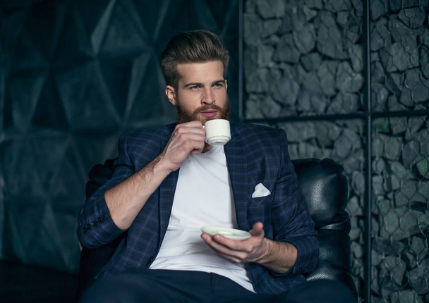 Бизнесмен с чашкой кофе сидит на современном фоне
. - Фото, изображение