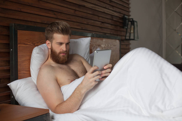 Sexy young man is lying in bed with tablet in hands. - Foto, afbeelding