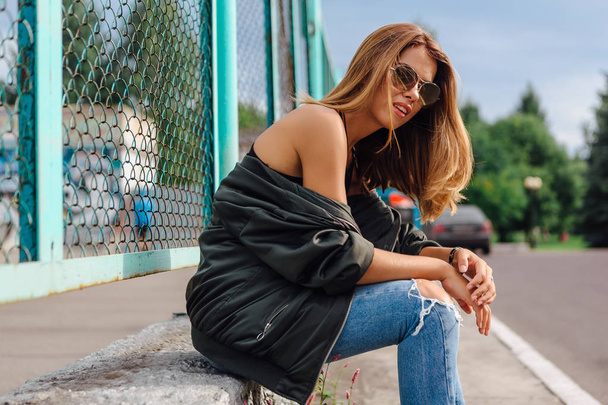 Fashion portrait of trendy young woman wearing sunglasses, jeans with halls and bomber jacket in the city. Copy space. - Фото, изображение